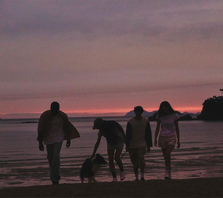 Silhouetten gehen am Strand bei Sonnenuntergang. Person streichelt Hund. Rosa und lila Farbtöne am Himmel spiegeln sich im Wasser.