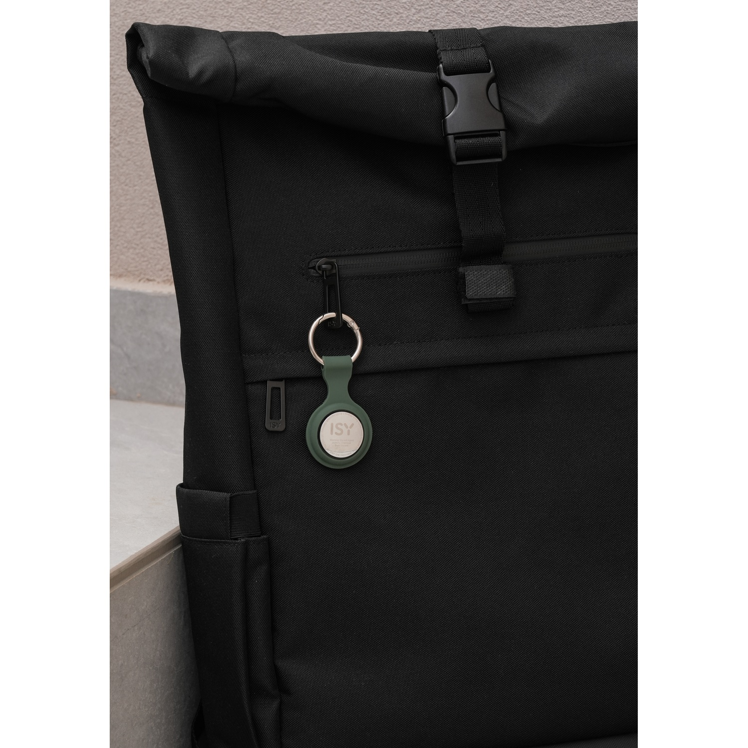 Zaino nero con chiusura arrotolabile e fibbia nera. Un portachiavi verde con un'etichetta rotonda pende dalla cerniera.