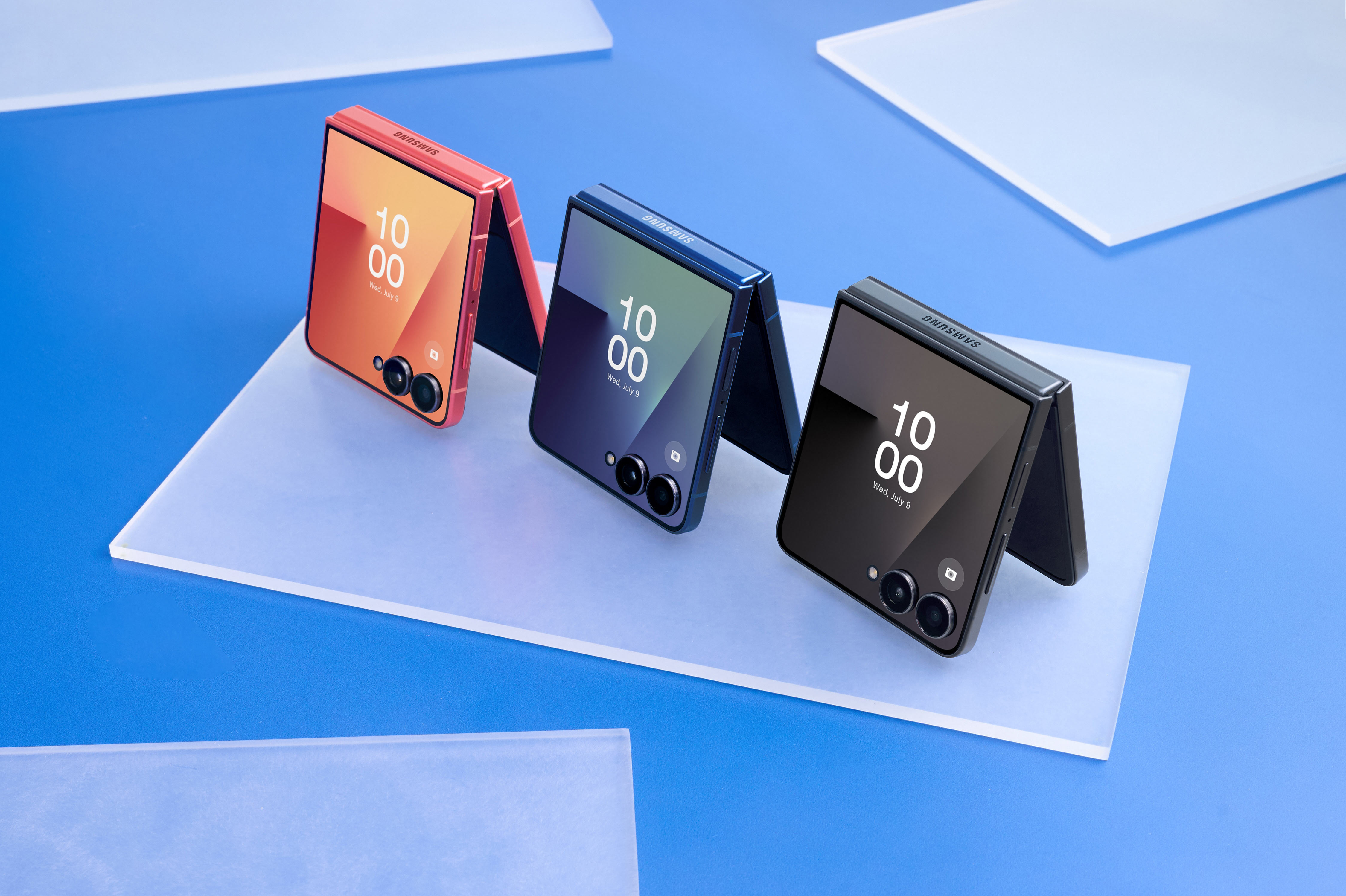 Trzy składane telefony Samsung w kolorze czerwonym, niebieskim i czarnym, wyświetlające godzinę i datę, na białej powierzchni, niebieskie tło.