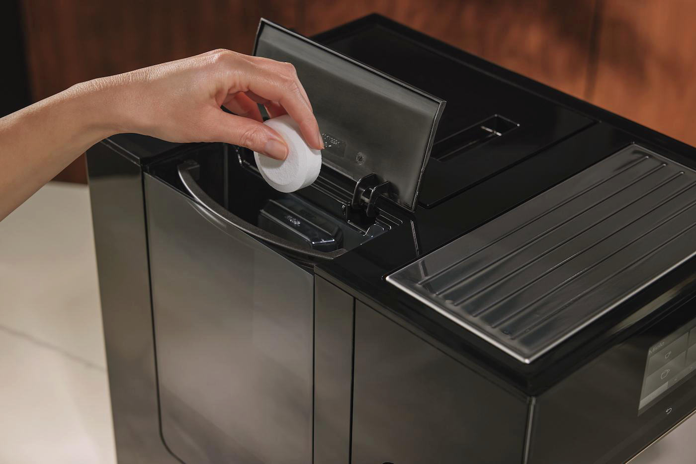 Eine Hand legt einen weißen Filter in eine schwarze Kaffeemaschine. Die Maschine hat ein Tablett und einen offenen Deckel.