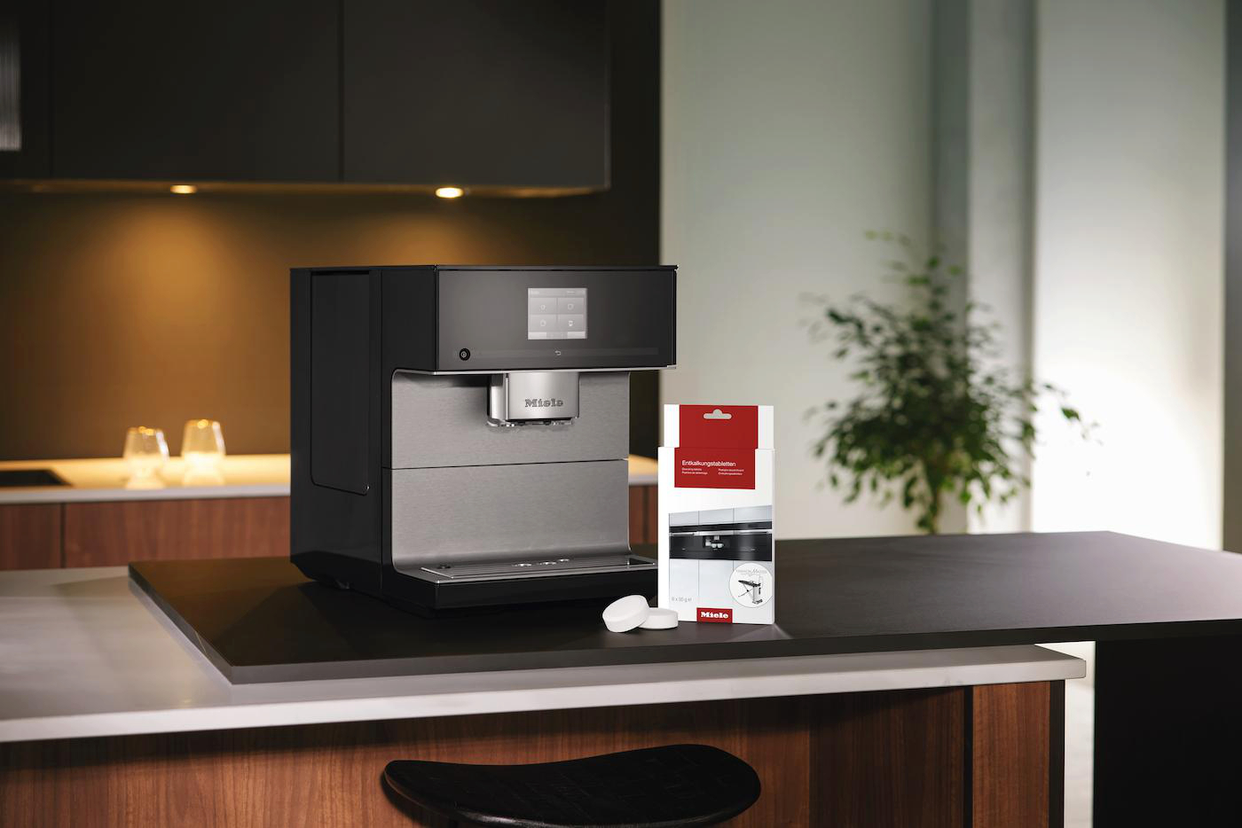 Auf einer Arbeitsplatte steht eine schwarze und silberne Miele-Kaffeemaschine mit Entkalkungstabletten und einer Produktverpackung.