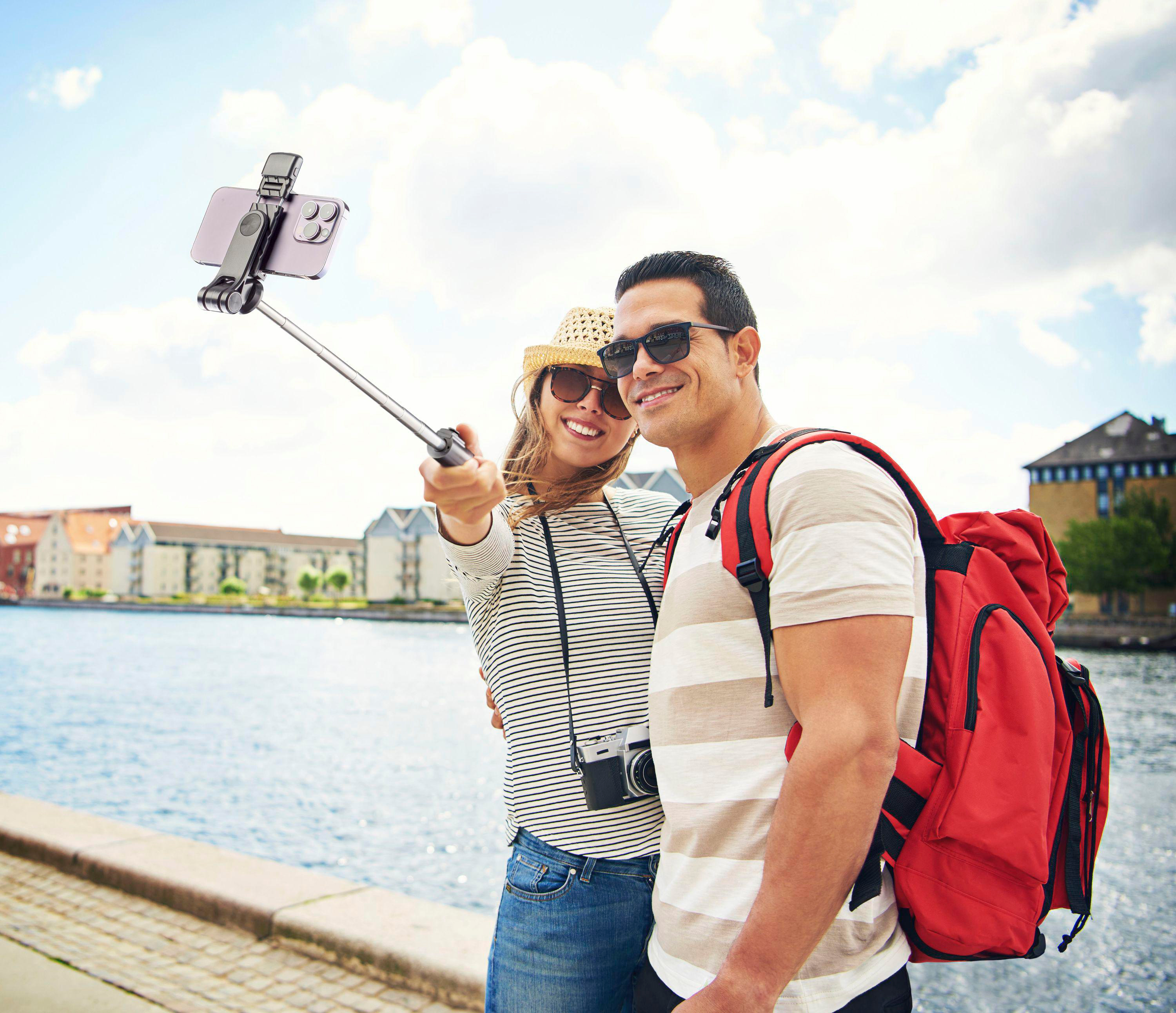 Ein Paar macht ein Selfie mit einem Telefon an einem Stock. Sie sind am Wasser. Gebäude im Hintergrund.