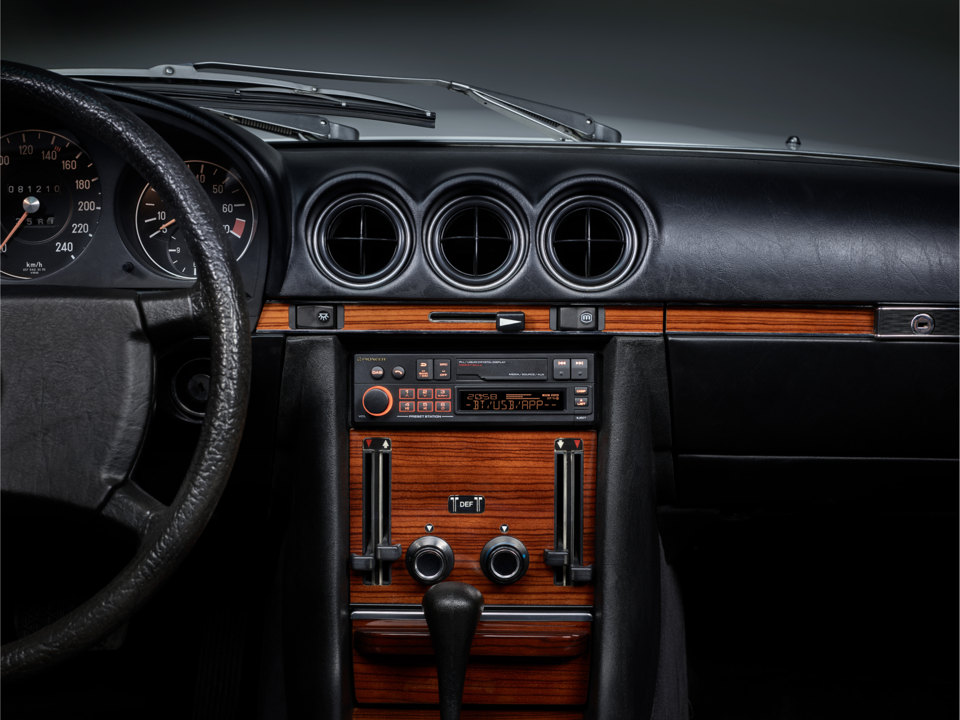 Tableau de bord d'une voiture classique, avec jauges, garnitures en bois, radio et bouches d'aération. Le volant est à gauche.
