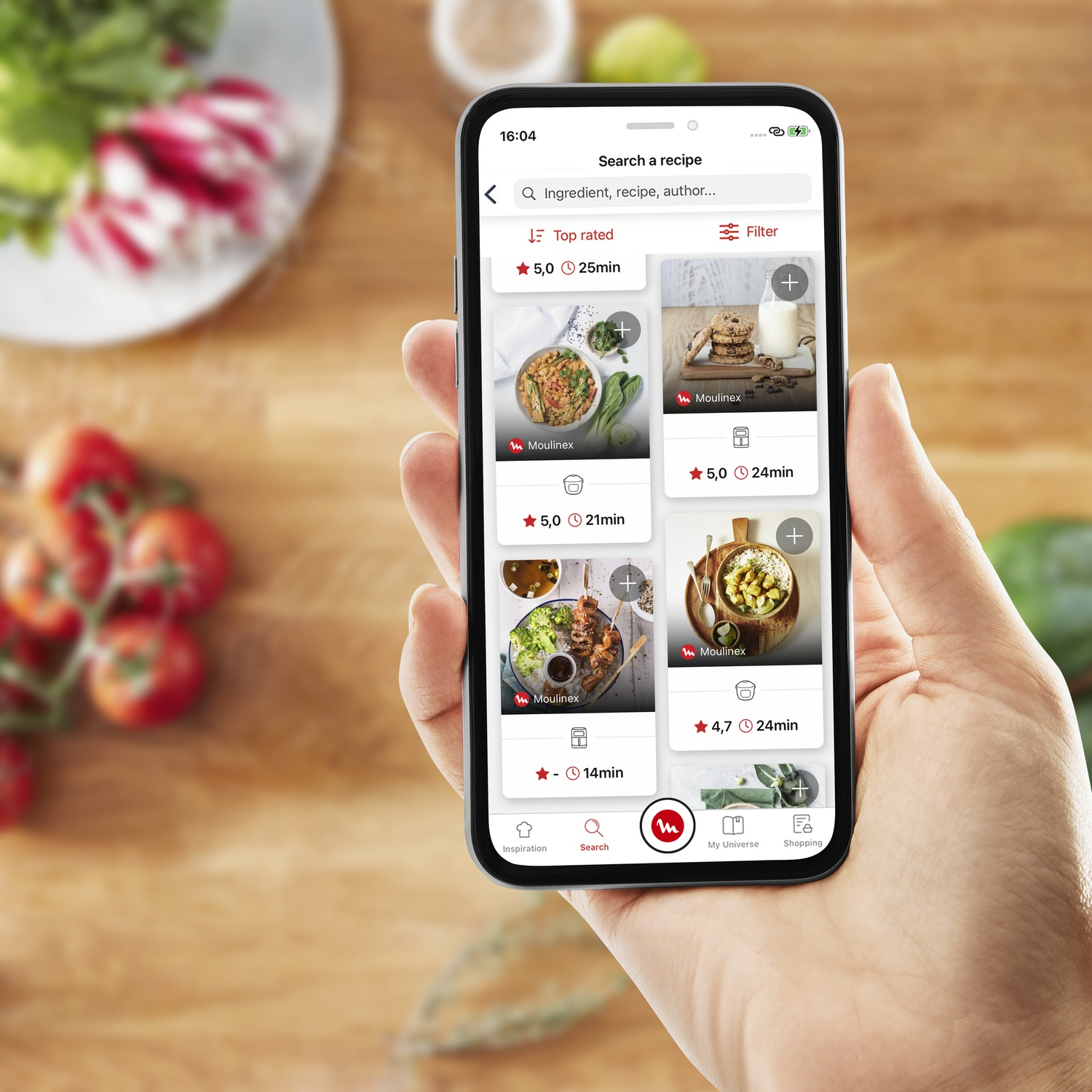 Een hand houdt een telefoon vast met een recepten-app. Vage achtergrond met groenten. App toont receptkaarten met beoordelingen en kooktijden.