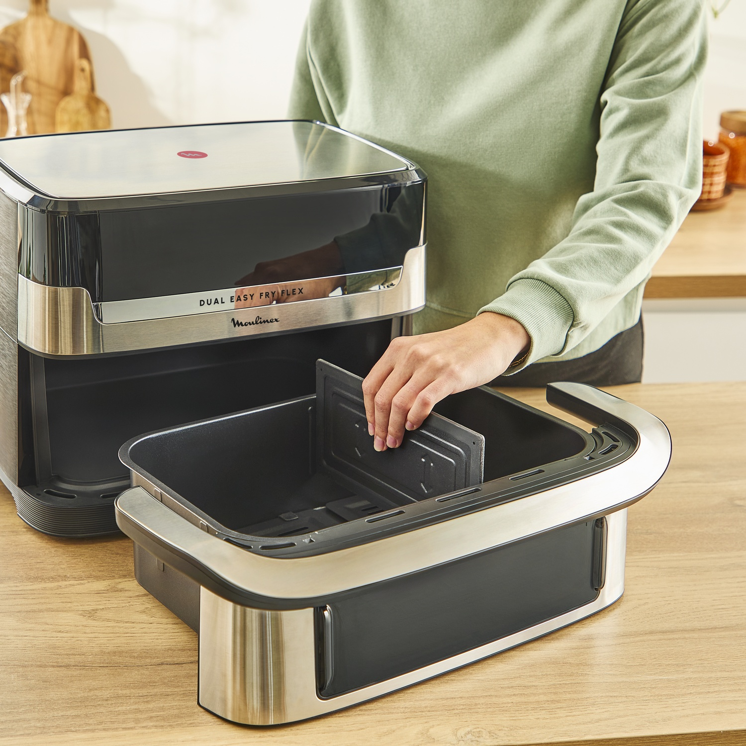 Een persoon plaatst een verdeler in een airfryer-mand. De airfryer is zwart en zilverkleurig en staat op een houten oppervlak.