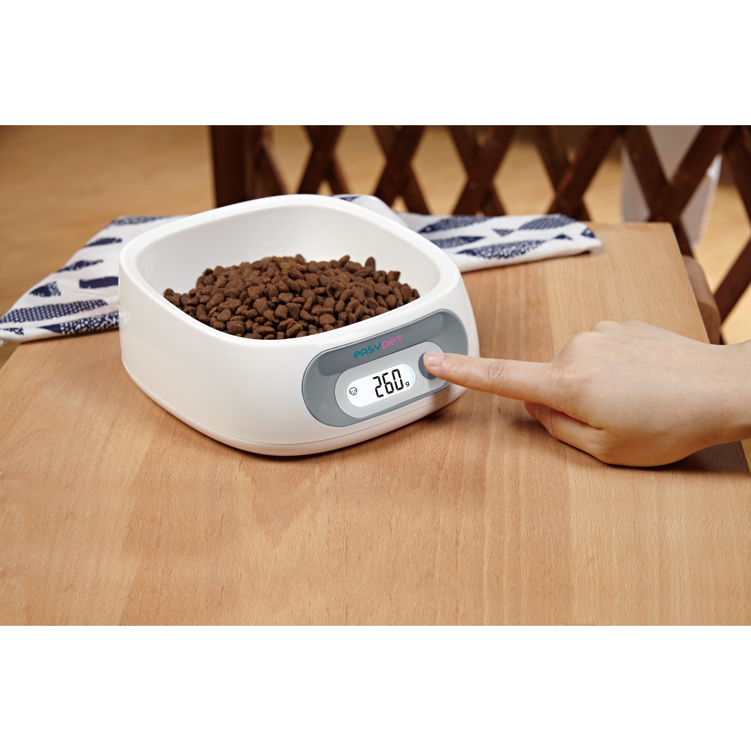 A white pet food bowl with brown kibble, a hand pressing a button, and a digital display showing 260g on a wooden surface.