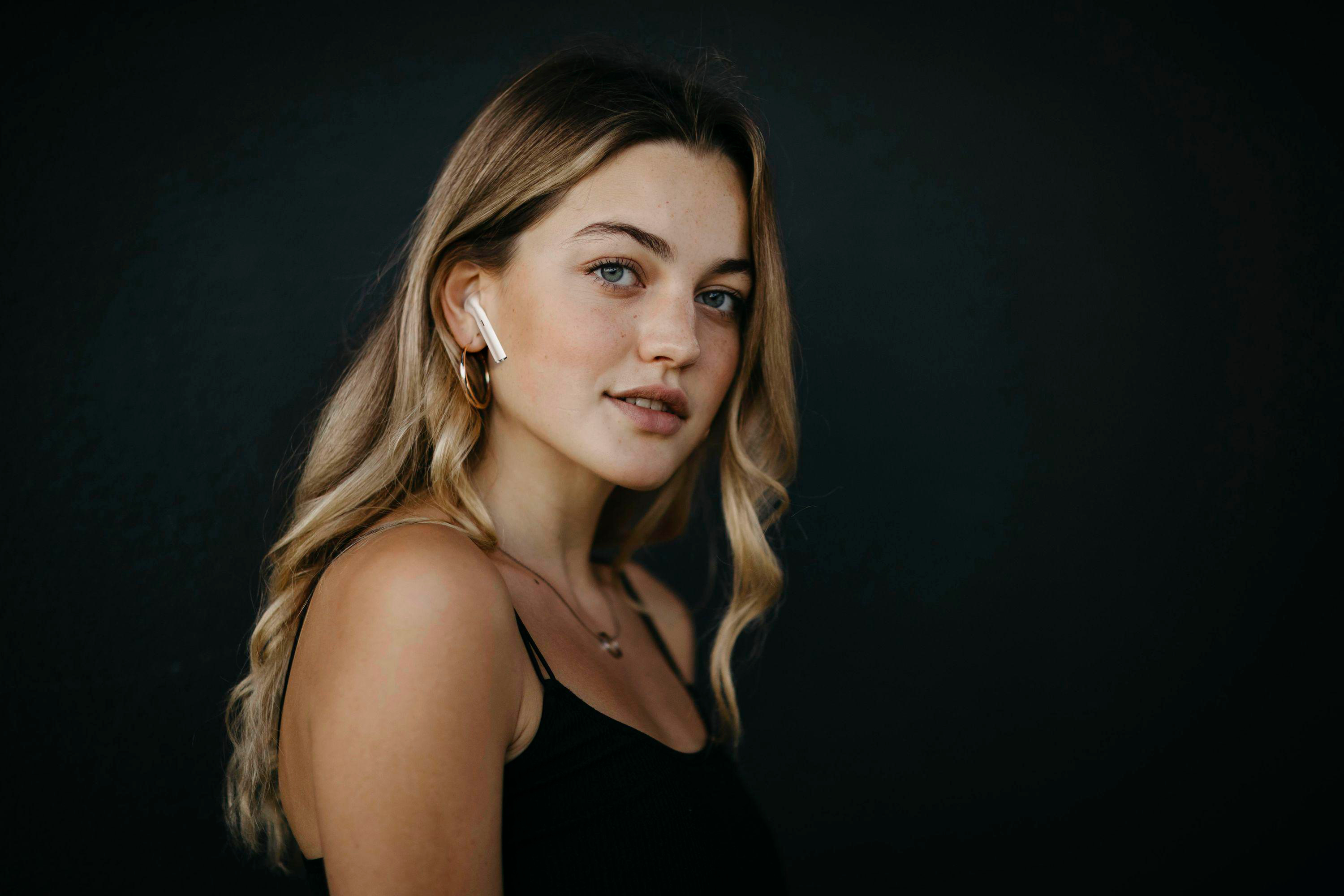 Eine junge Frau mit blonden Haaren und blauen Augen, trägt ein schwarzes Top und einen drahtlosen Ohrhörer, lächelt.