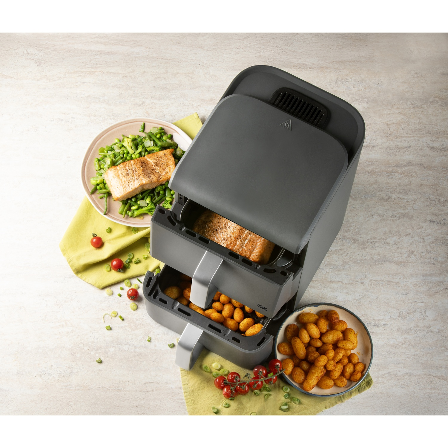 Un airfryer avec de la nourriture à l'intérieur, à côté d'une assiette avec du saumon et des légumes, et un bol avec des pommes de terre frites. Fond : table en pierre.
