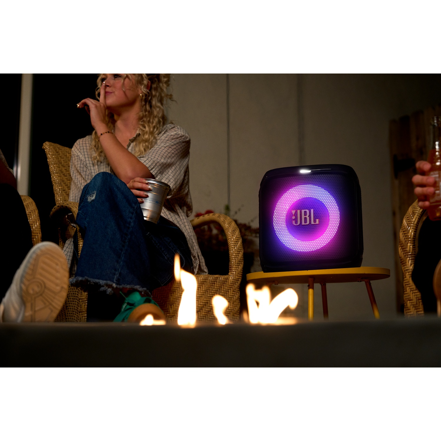 People around a fire pit with a JBL speaker on a yellow table. Woman holds a drink. Flames in the foreground, speaker in the middle.
