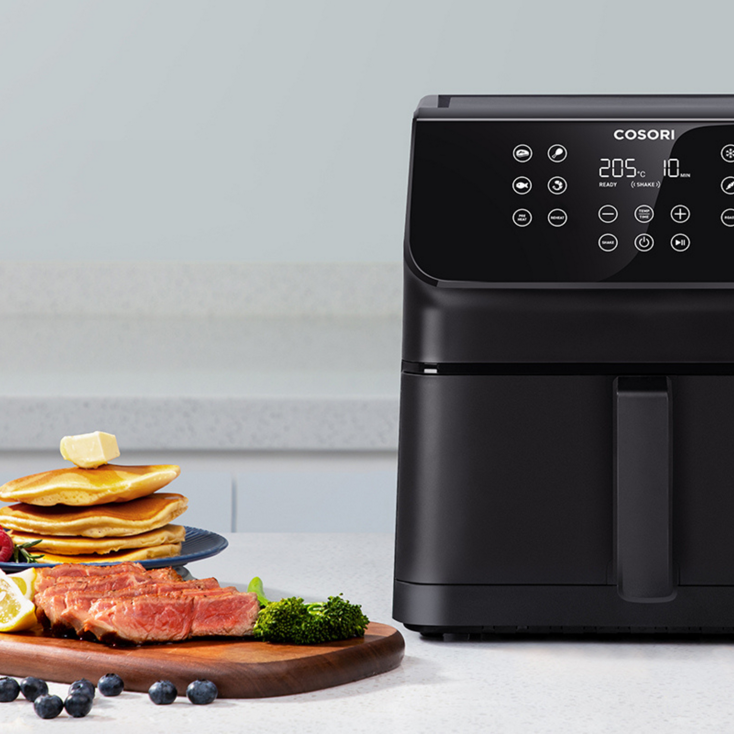 Freidora de aire negra con alimentos como bistec y panqueques. Tiene una pantalla digital y está sobre una encimera blanca.