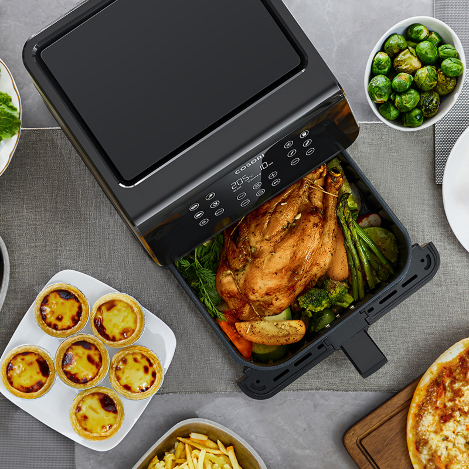 Una freidora de aire con pollo asado y verduras. También tiene tartas, coles de Bruselas, patatas fritas y pizza en la mesa.