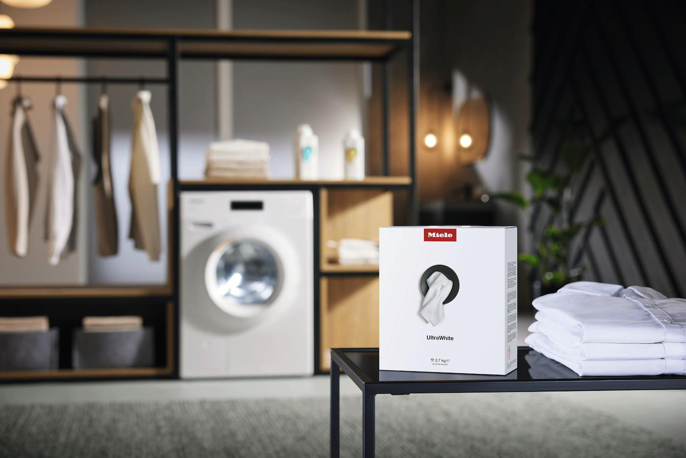 Ein Waschraum mit Waschmaschine, Kleiderständer und einer Schachtel Miele UltraWhite Waschmittel auf einem Tisch.