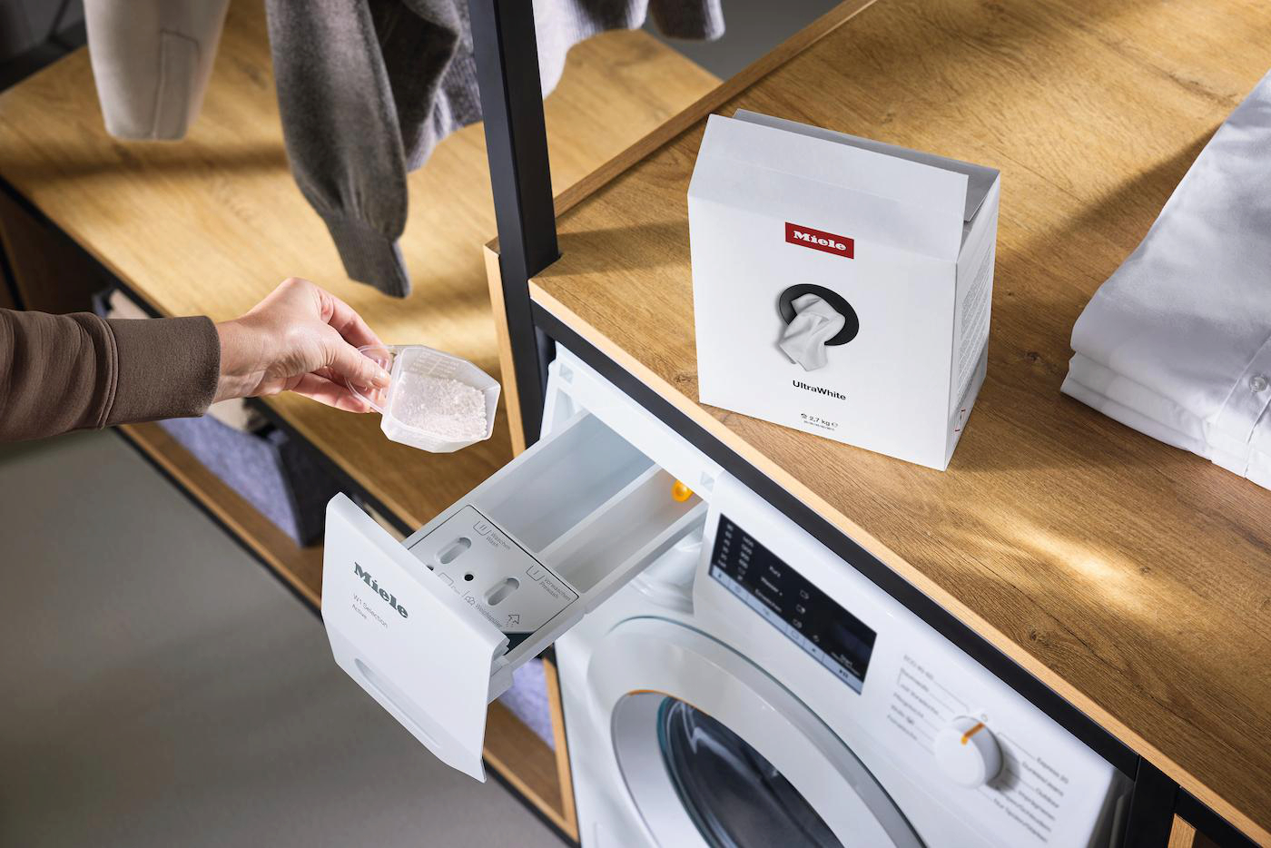 Eine Person gießt Waschmittel in eine Waschmaschinenschublade. Eine Schachtel UltraWhite Waschmittel und Kleidung sind in der Nähe.