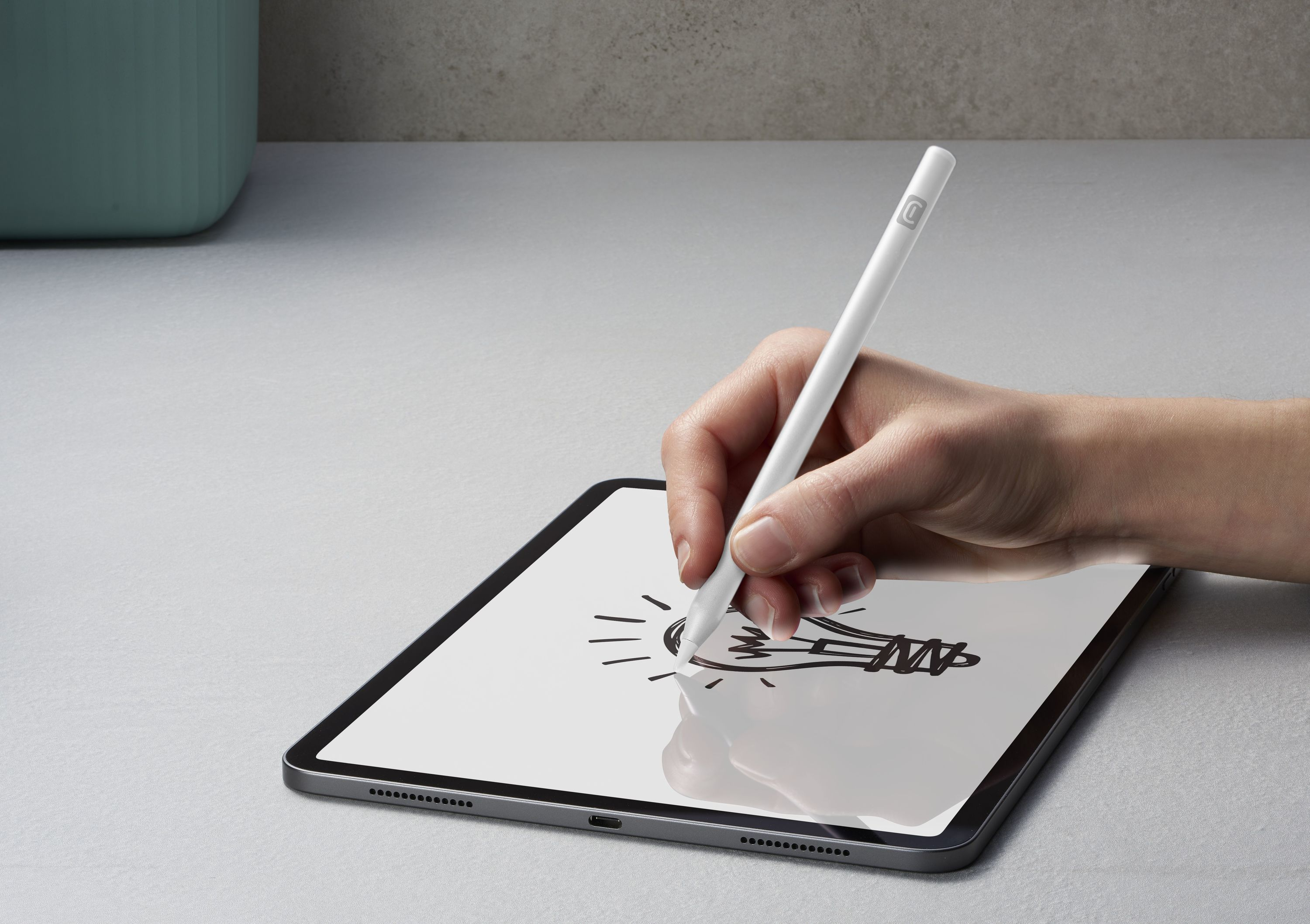 Beyaz bir kalem tutan el, bir tablet üzerine ampul çiziyor. Tablet açık gri bir yüzeyde.