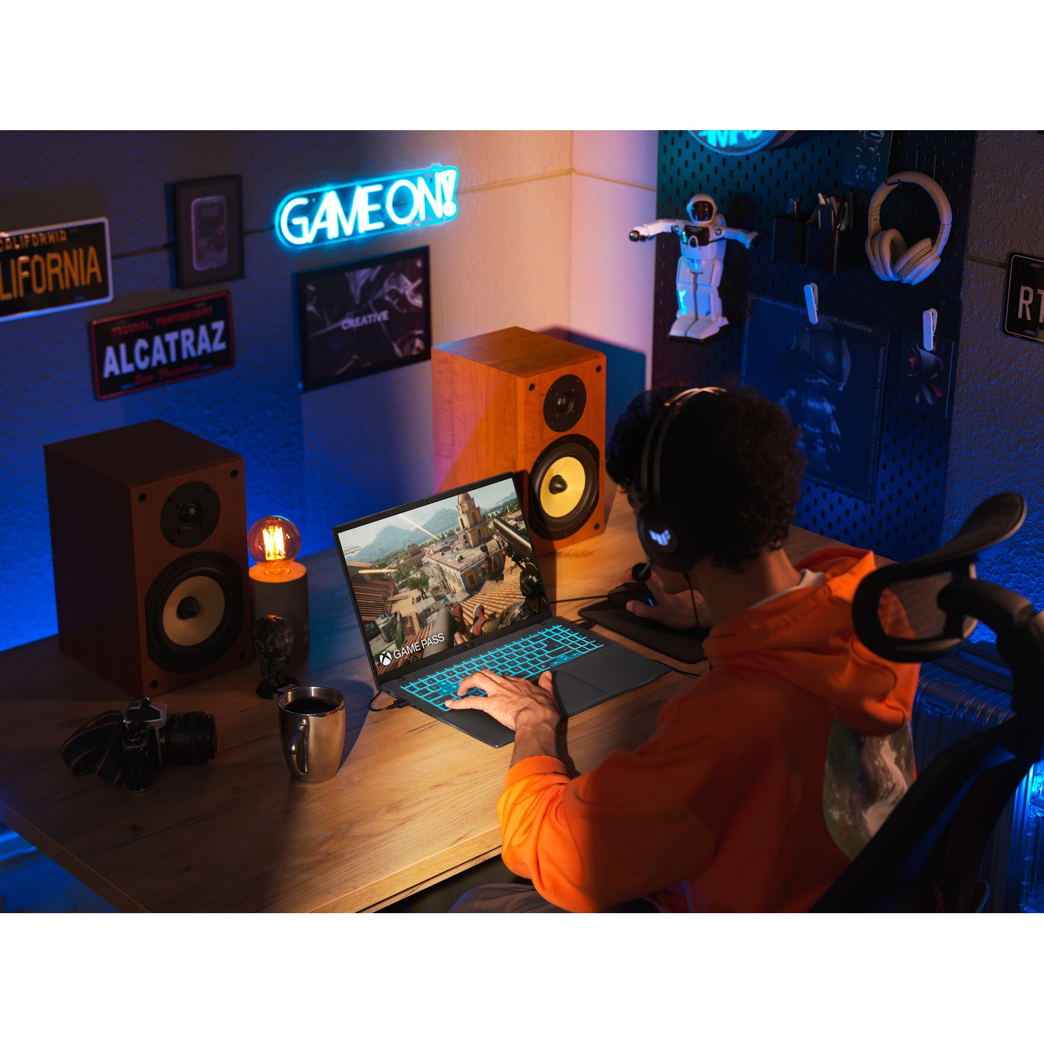 Una persona sta giocando su un laptop. Ci sono altoparlanti, un robot e un'insegna al neon che dice GAME ON!. La persona indossa le cuffie.