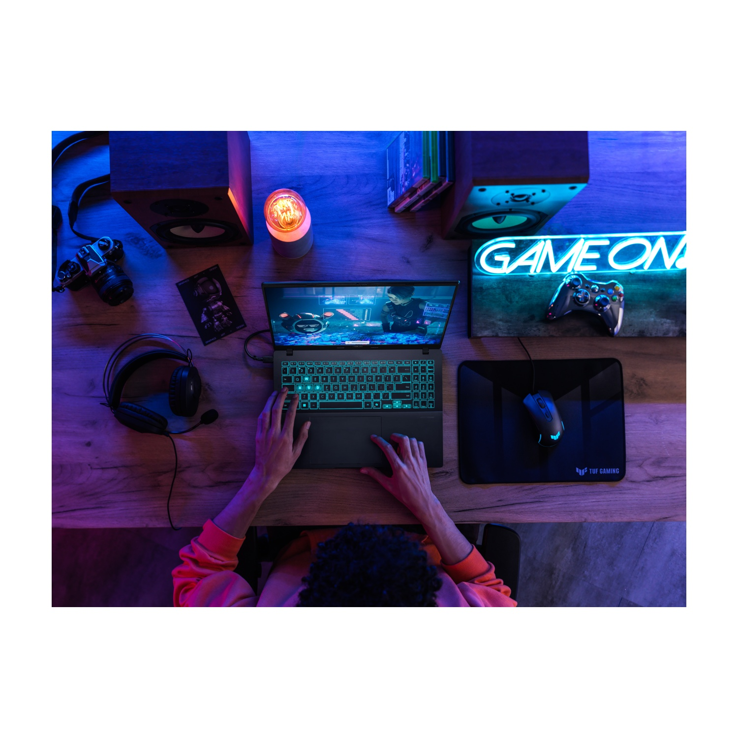 Una persona usa un laptop, con un mouse e cuffie su una scrivania di legno. Un cartello 'GAME ON' e altoparlanti sullo sfondo.