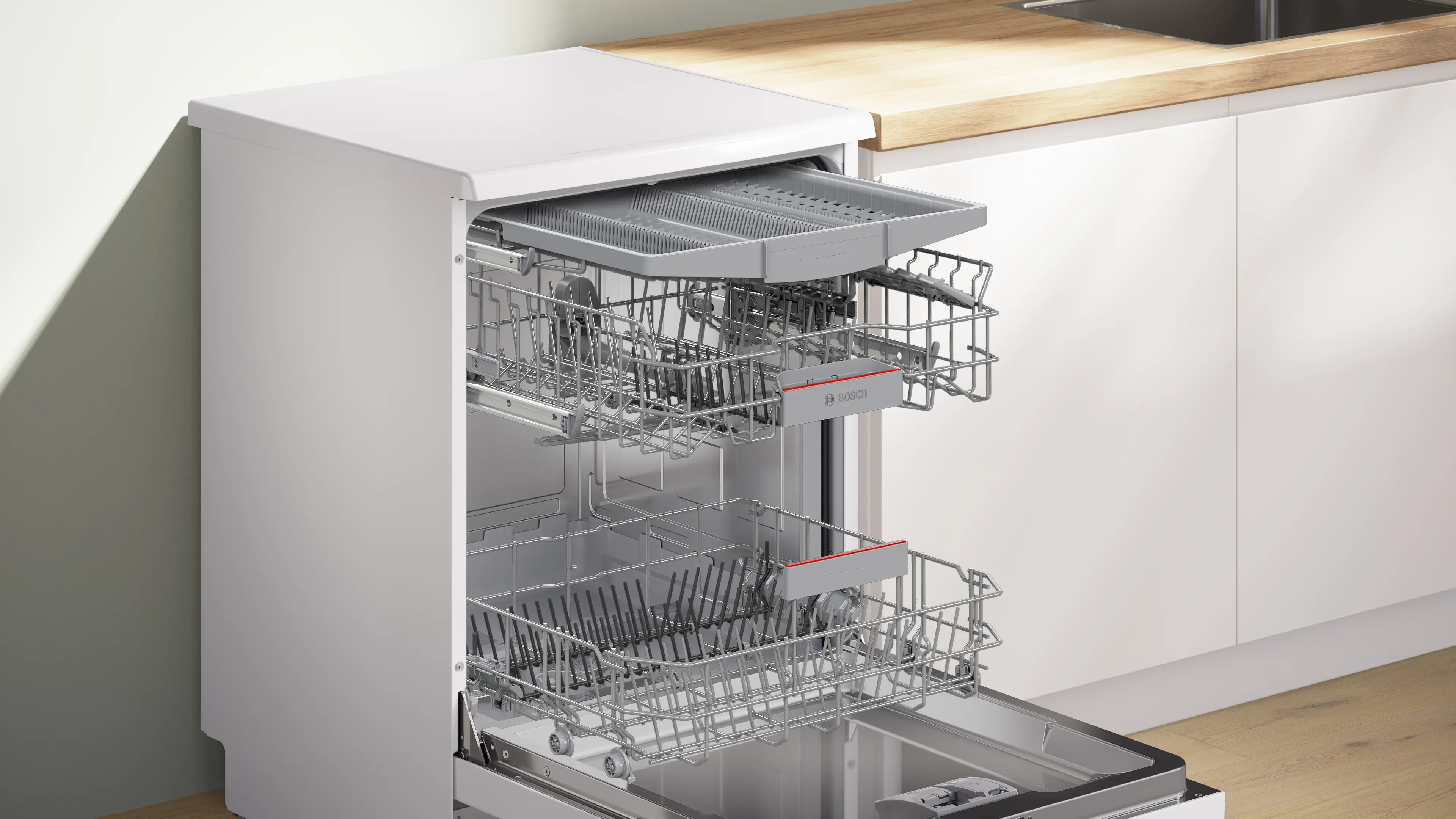 Lavavajillas blanco abierto con dos rejillas, una bandeja para utensilios y un interior de acero inoxidable. La encimera de la cocina es de madera.