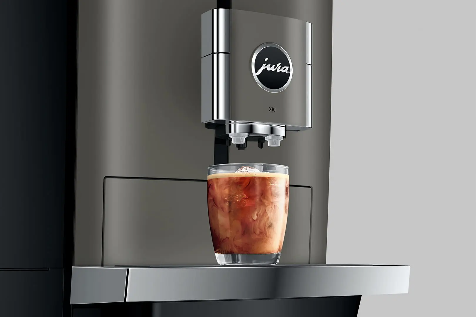 Una macchina da caffè che eroga caffè freddo in un bicchiere. La macchina è grigia, con un logo 'Jura'. Lo sfondo è grigio chiaro.