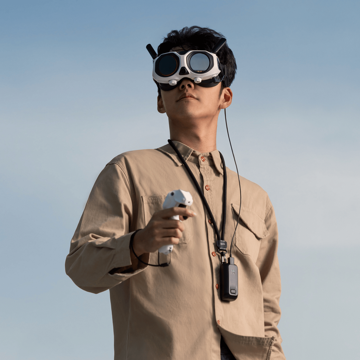 Un homme portant un masque VR et tenant une manette. Il porte une chemise beige et une sangle de cou. Ciel bleu en arrière-plan.
