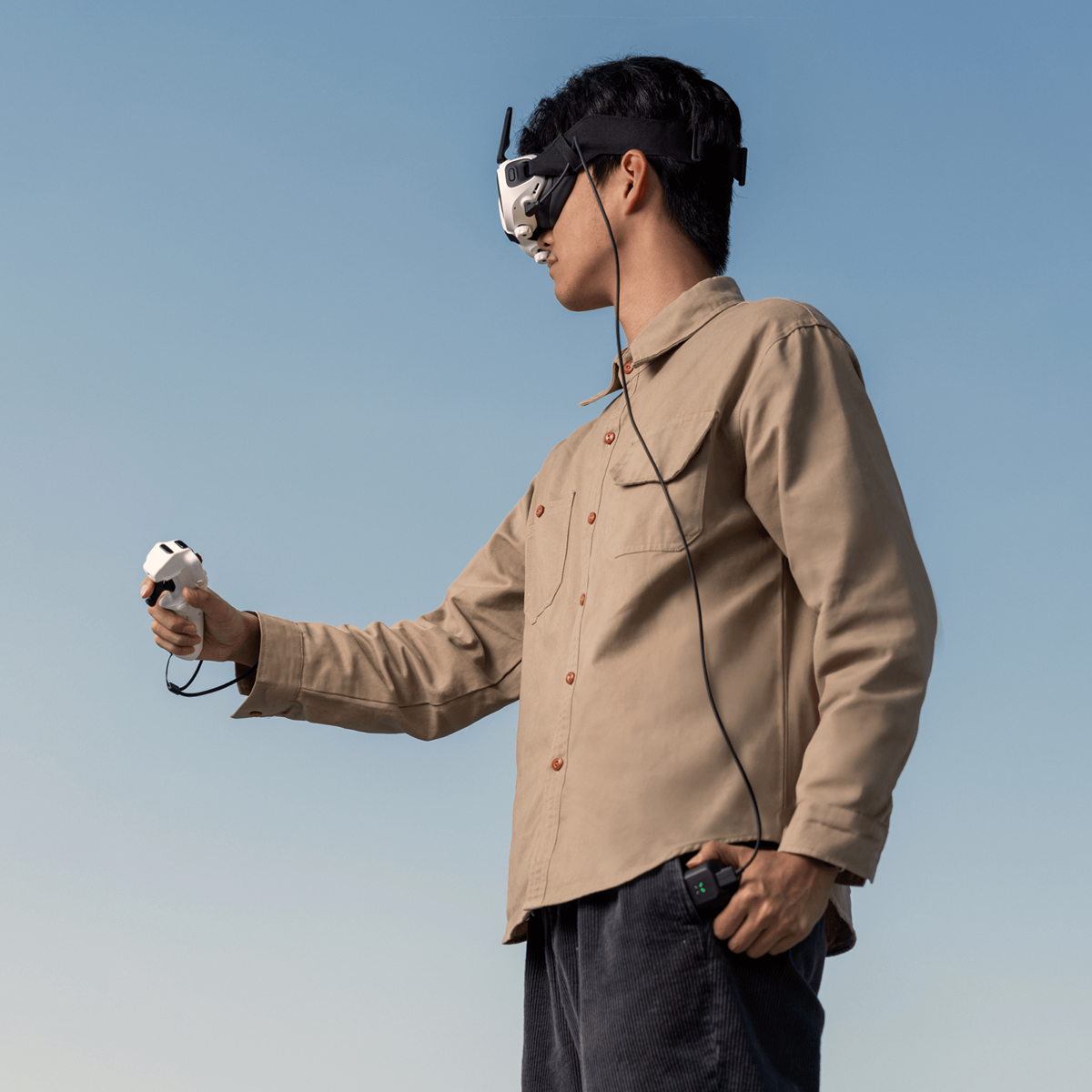 Un homme portant des lunettes VR et tenant une manette. Il porte une chemise beige et un pantalon sombre contre ciel bleu.