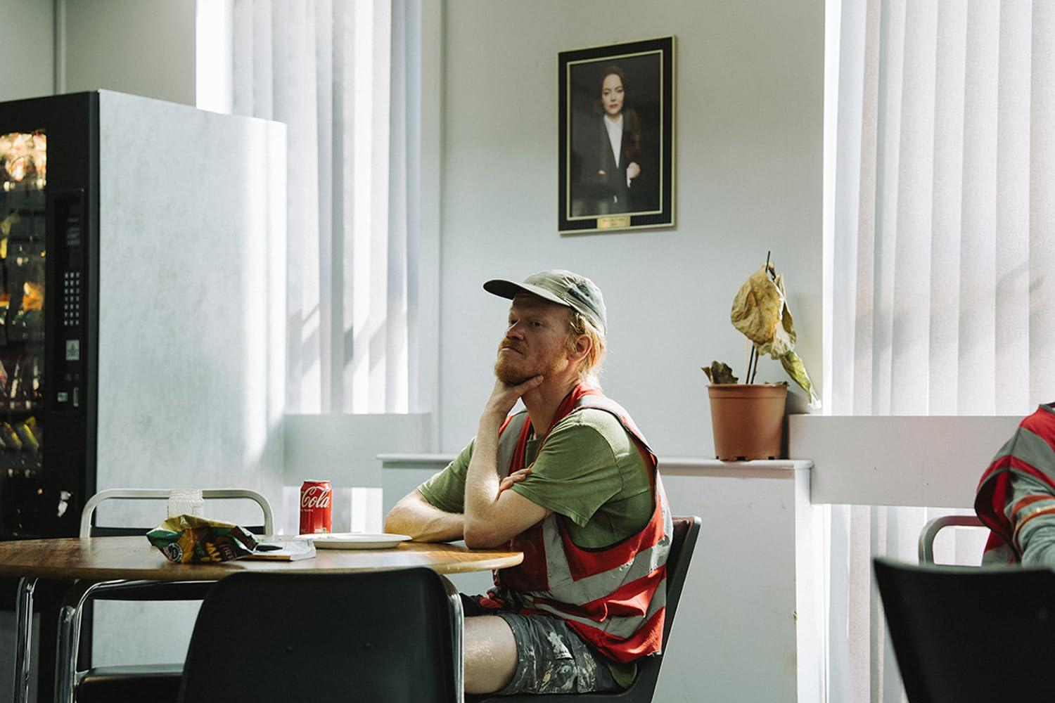 Ein Mann sitzt an einem Tisch mit Soda und Chips. Er trägt eine Kappe und Weste. Ein Porträt hängt an der Wand, und eine Pflanze steht in der Nähe.