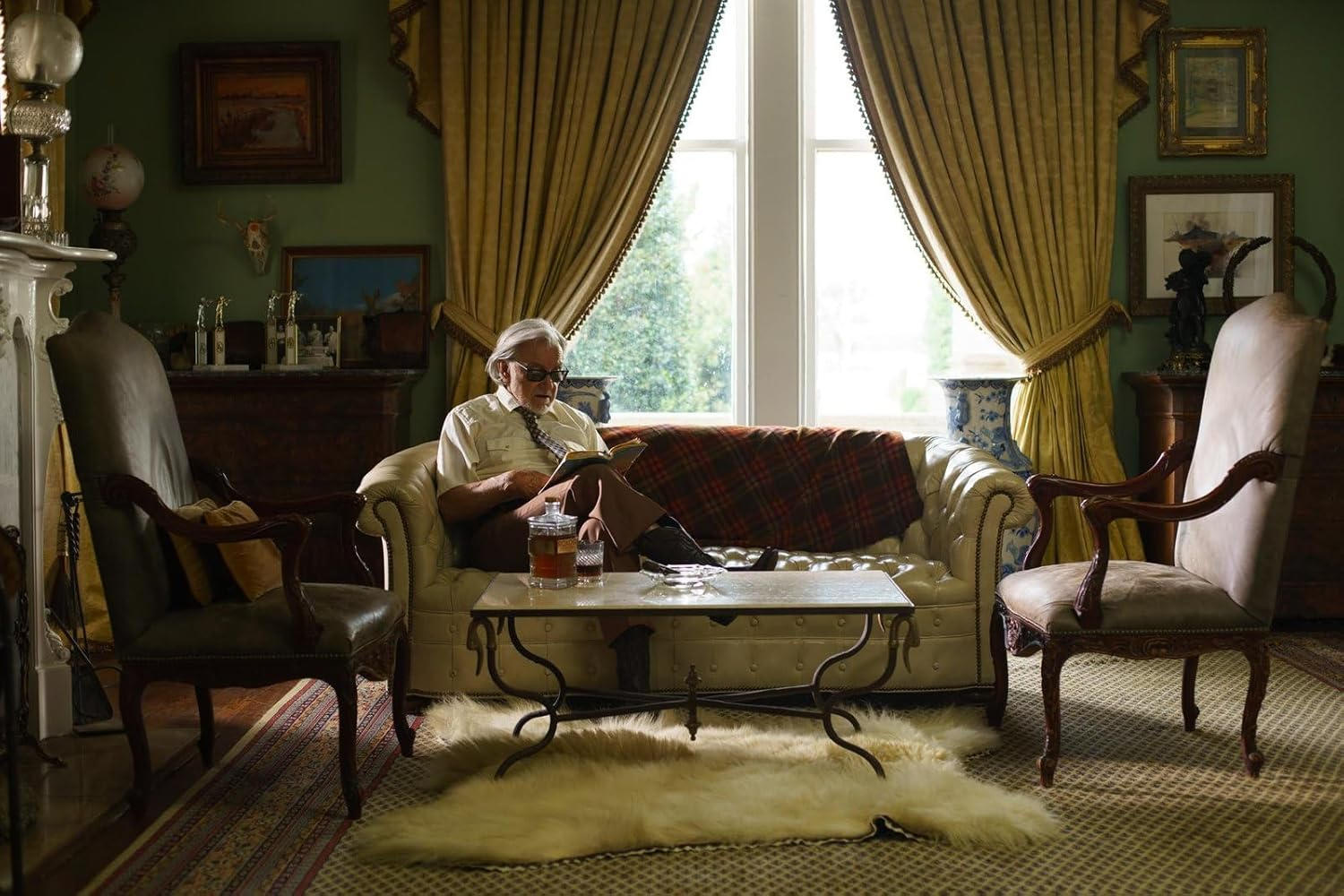 Ein älterer Mann liest in einem schicken Wohnzimmer. Er sitzt auf einem weißen Sofa mit einer karierten Decke. Der Raum hat Vorhänge und einen Teppich.