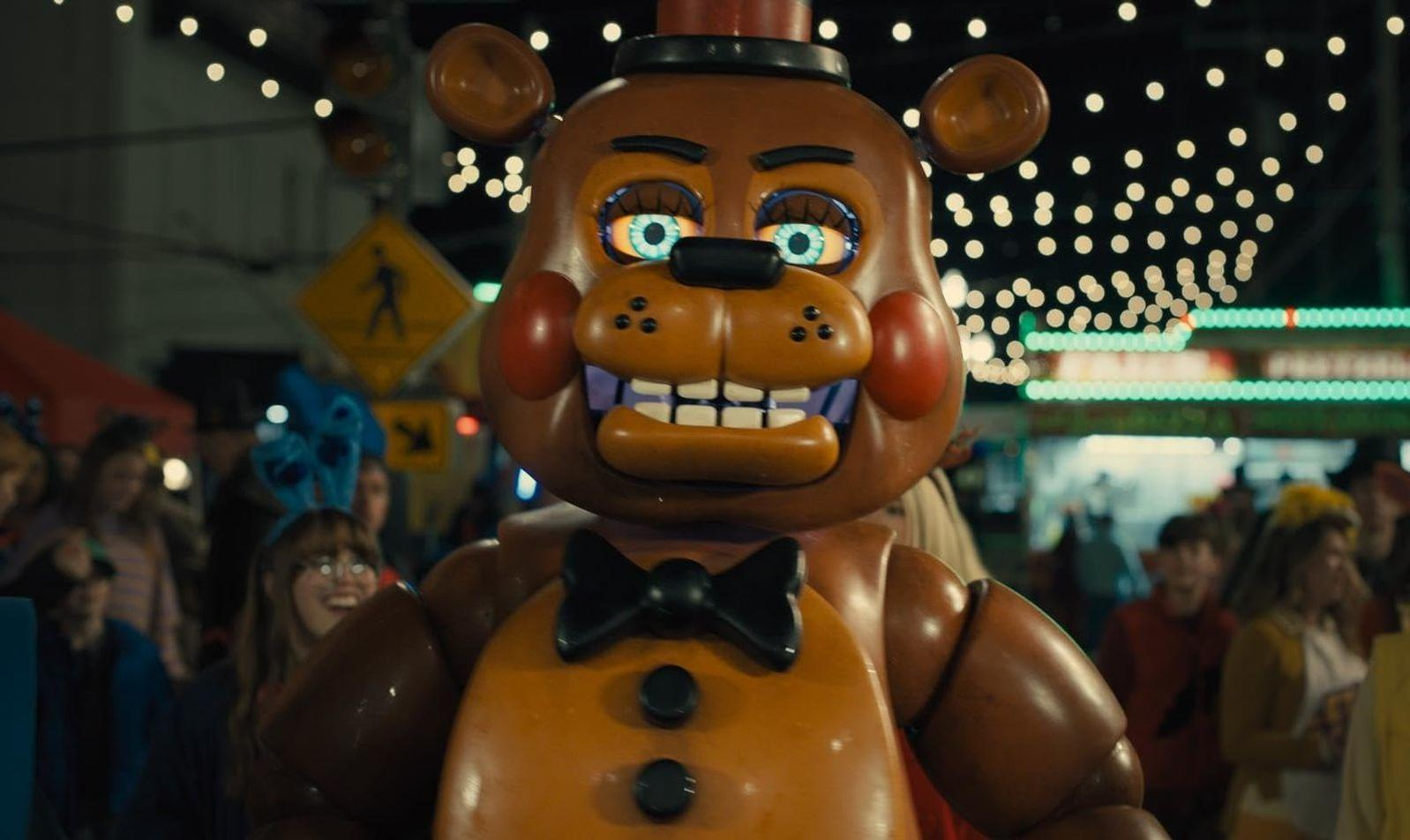 Ein großer Freddy Fazbear-Animatronic steht auf einem Jahrmarkt. Im Hintergrund sind Menschen. Lichter und Schilder sind sichtbar.