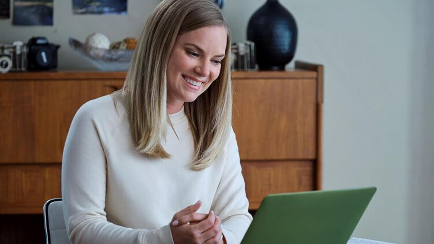 Eine Frau mit blonden Haaren lächelt auf einen grünen Laptop. Sie trägt einen weißen Pullover. Holzschrank und Dekoration im Hintergrund.