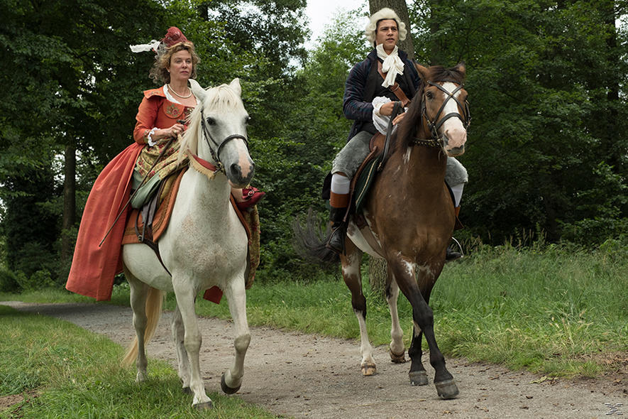 Zwei Personen in historischer Kleidung reiten auf einem Pfad. Bäume säumen den Weg. Ein Pferd ist weiß, das andere braun.