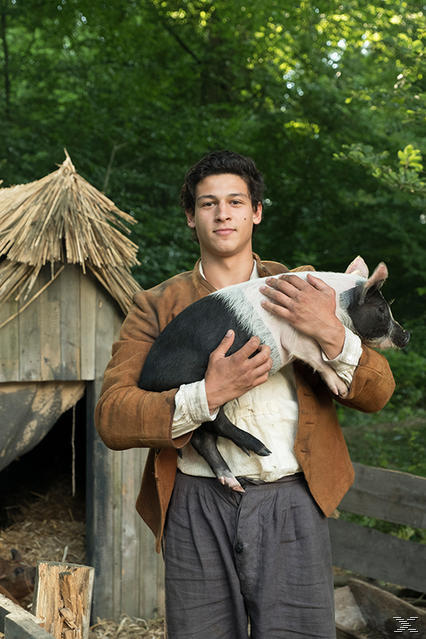 Ein junger Mann hält ein schwarz-weißes Ferkel. Er trägt eine braune Jacke und graue Hose. Im Hintergrund ist ein Schweinestall aus Holz.