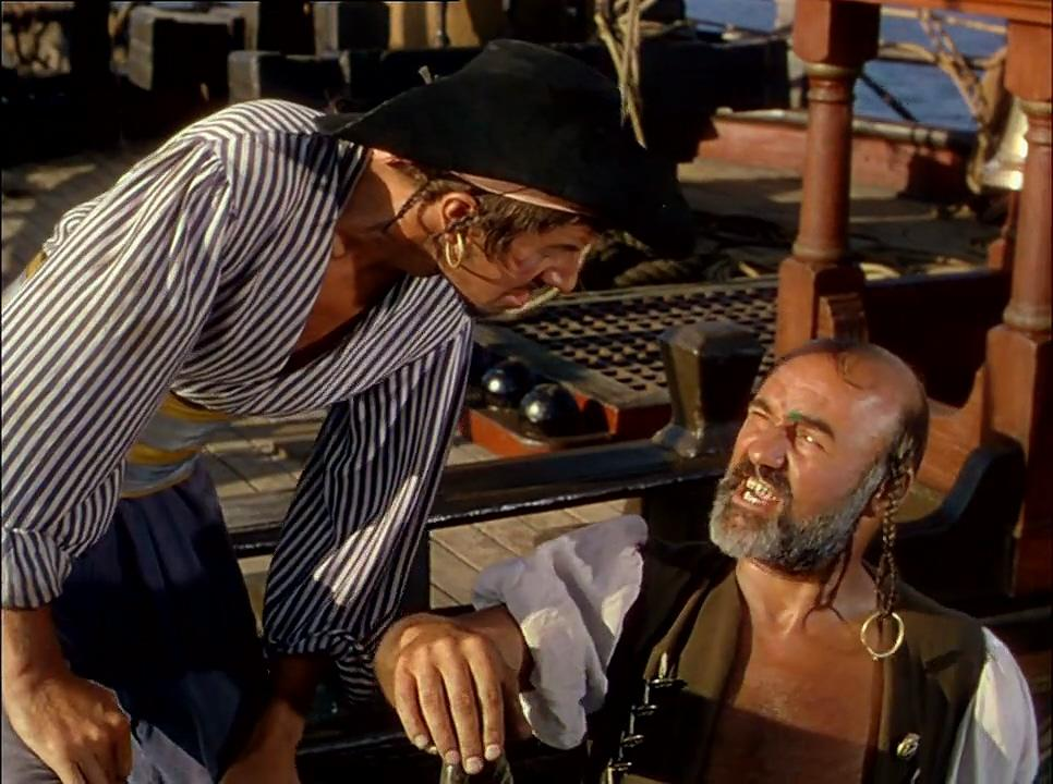 Zwei Piraten auf einem Schiff. Einer im gestreiften Hemd beugt sich über den anderen, der mit Bart, Ohrring und weißem Tuch auf dem Deck liegt.