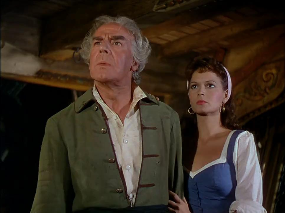 Ein älterer Mann in einer grünen Jacke und eine Frau in einem blauen Kleid schauen nach oben. Sie sind auf einem Schiff. Der Hintergrund ist aus Holz.