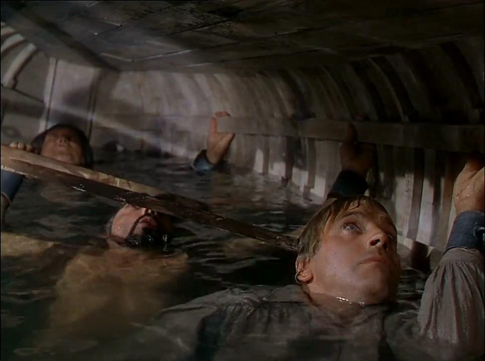 Drei Männer sind in einem Holzbauwerk unter Wasser. Sie kämpfen darum, über Wasser zu bleiben und blicken nach oben.