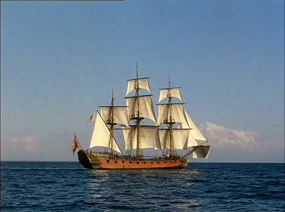 Ein dreimastiges Segelschiff mit weißen Segeln auf blauem Wasser unter klarem Himmel. Das Schiff hat einen roten Rumpf.