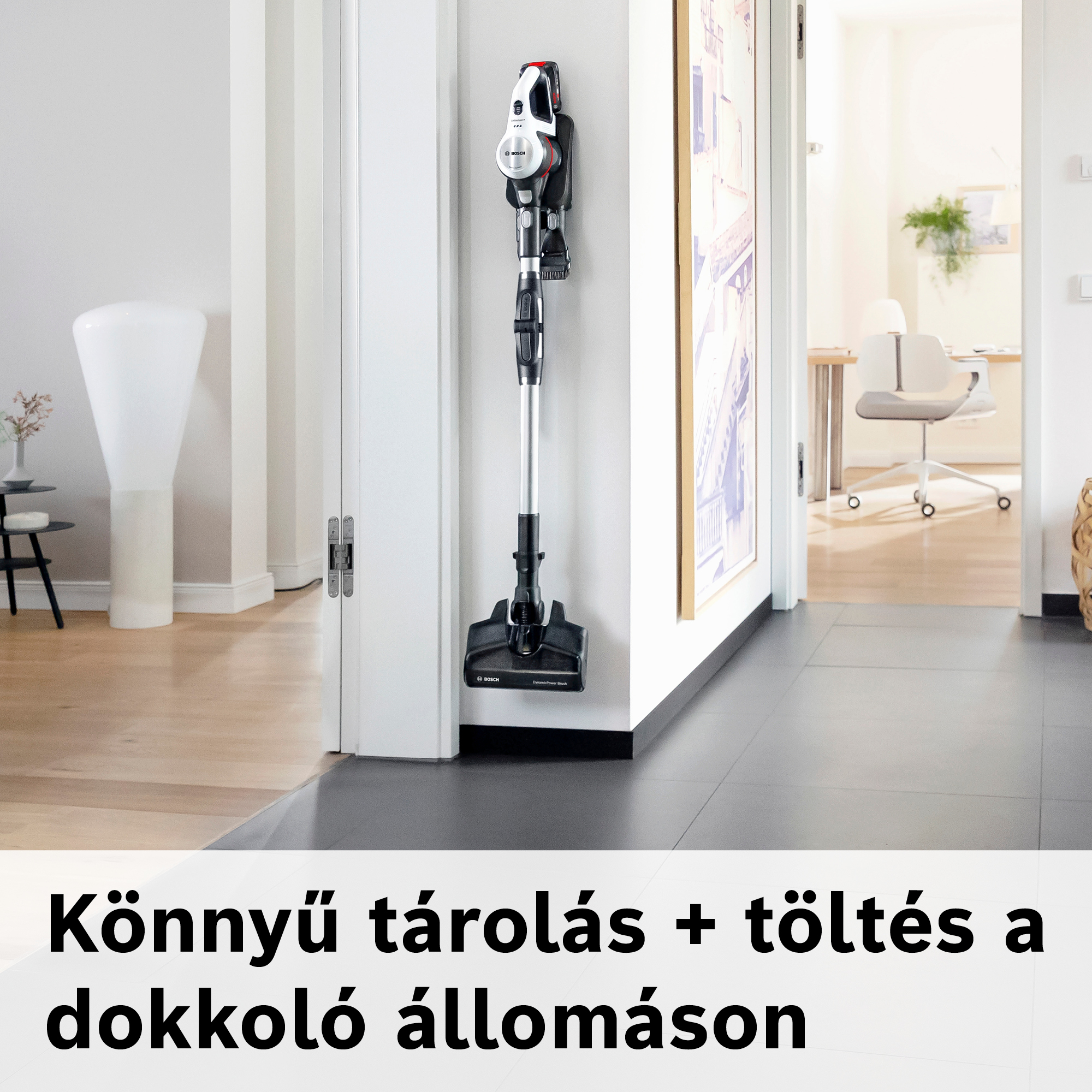 Egy vezeték nélküli porszívó van a falra szerelve. Fekete, fehér és ezüst. Egy ajtó és egy szoba látható.