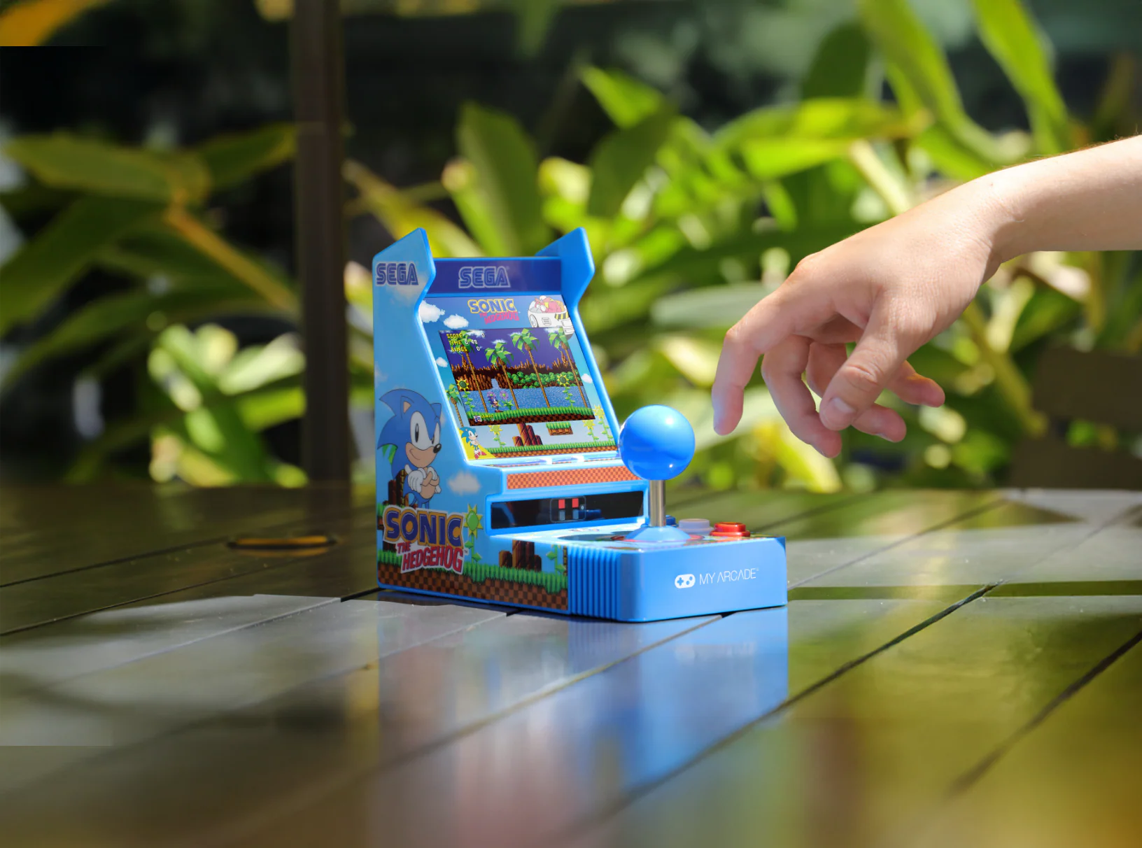 Una mano se acerca a una pequeña máquina recreativa azul de Sonic the Hedgehog. El fondo muestra follaje verde y una mesa oscura.