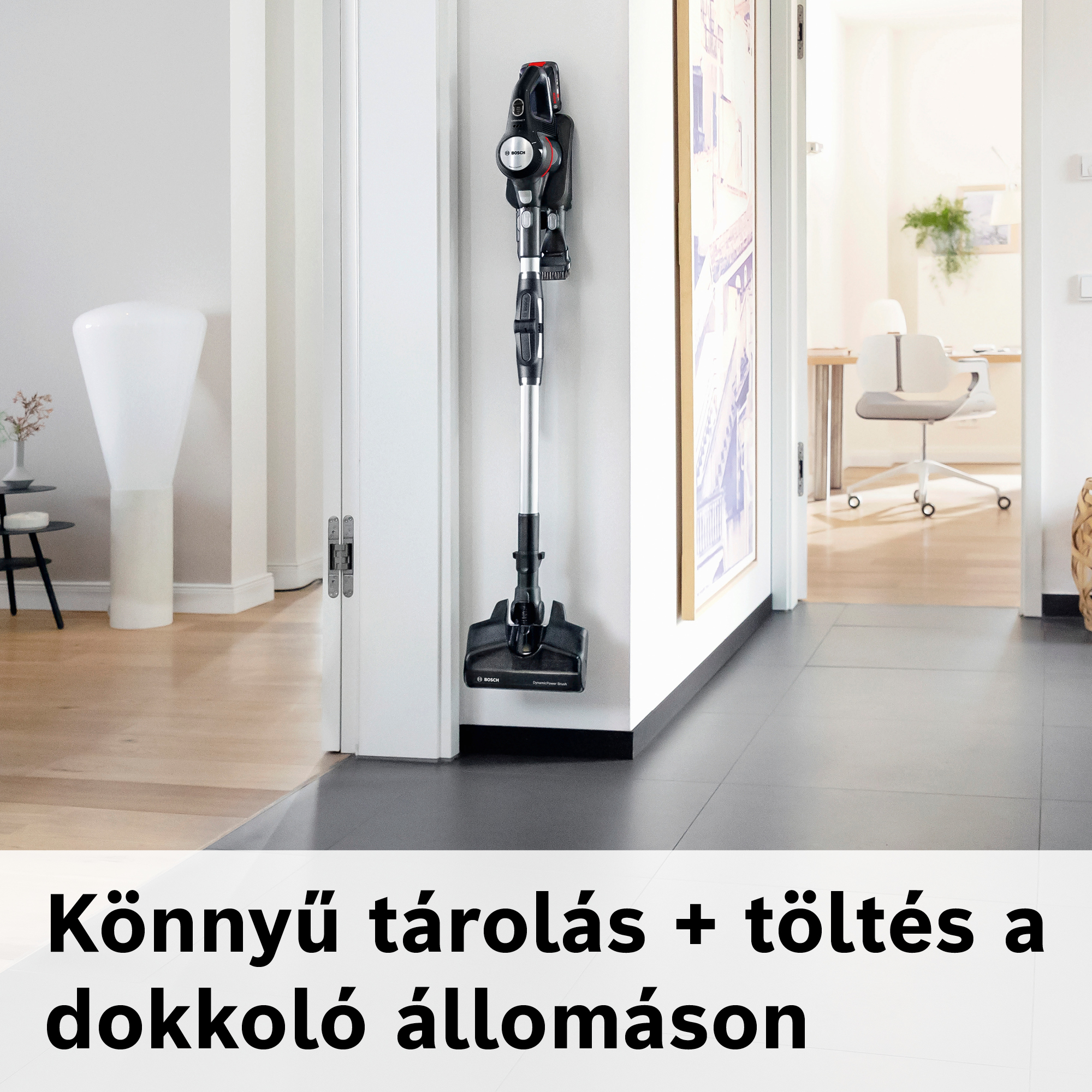 Egy fekete-ezüst vezeték nélküli porszívó fehér falra van szerelve. Az alatta lévő szöveg a következő: Könnyű tárolás + töltőállomás.