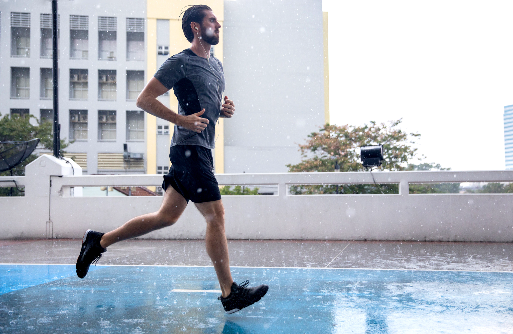 Un homme court sous la pluie sur une surface bleue. Il porte un short noir, une chemise grise et des chaussures noires. Des bâtiments sont en arrière-plan.