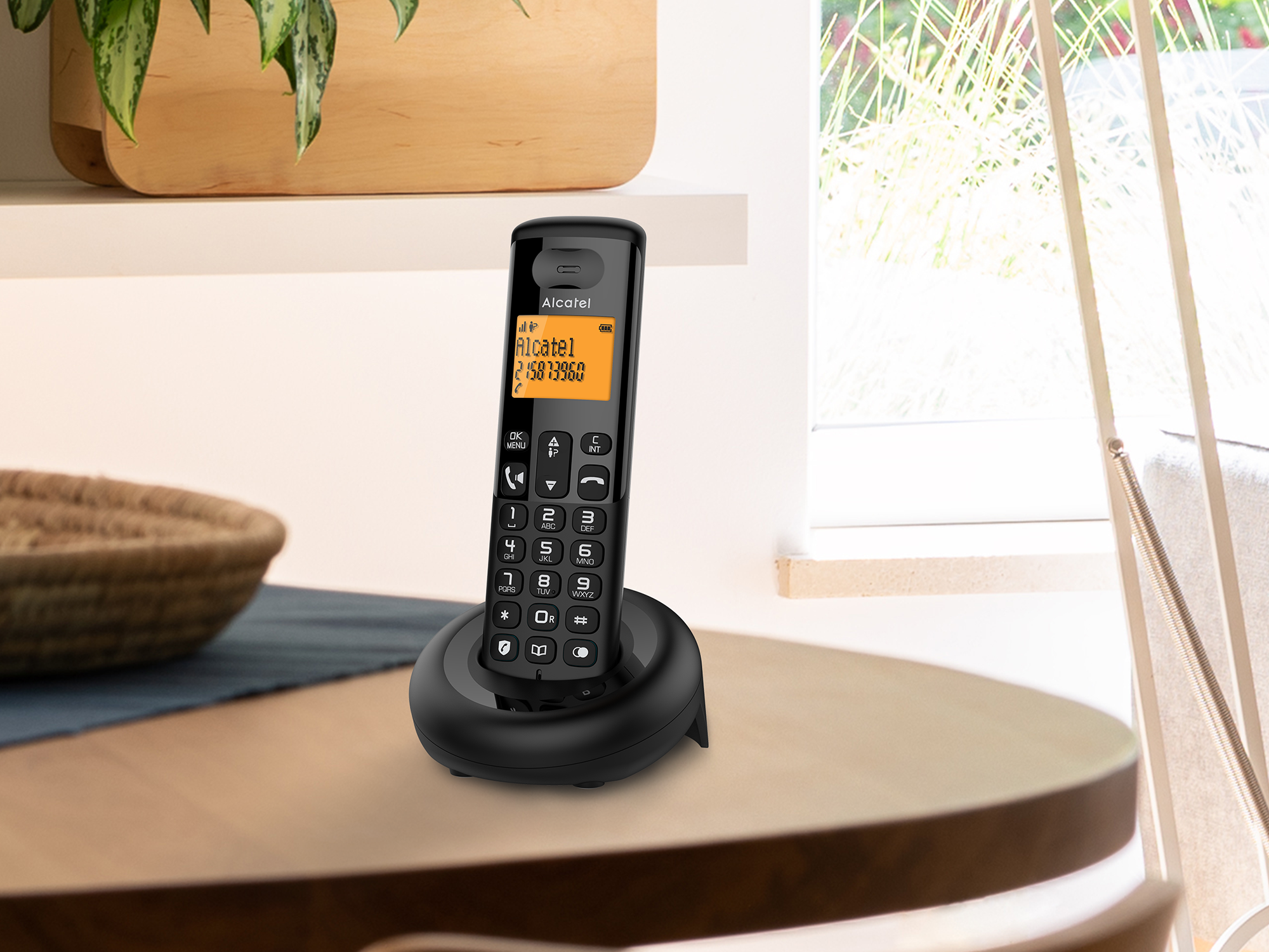 Egy fekete Alcatel telefon egy faasztalon. Egy kosár és egy növény a háttérben. Jobbra egy ablak látható.