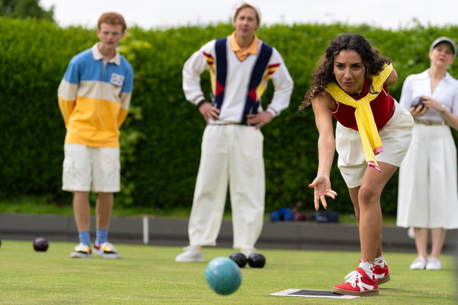Eine Frau in einem roten Oberteil wirft einen Ball auf einen grünen Rasen. Andere schauen zu. Bälle, grüne Hecke und weiße Shorts sind sichtbar.