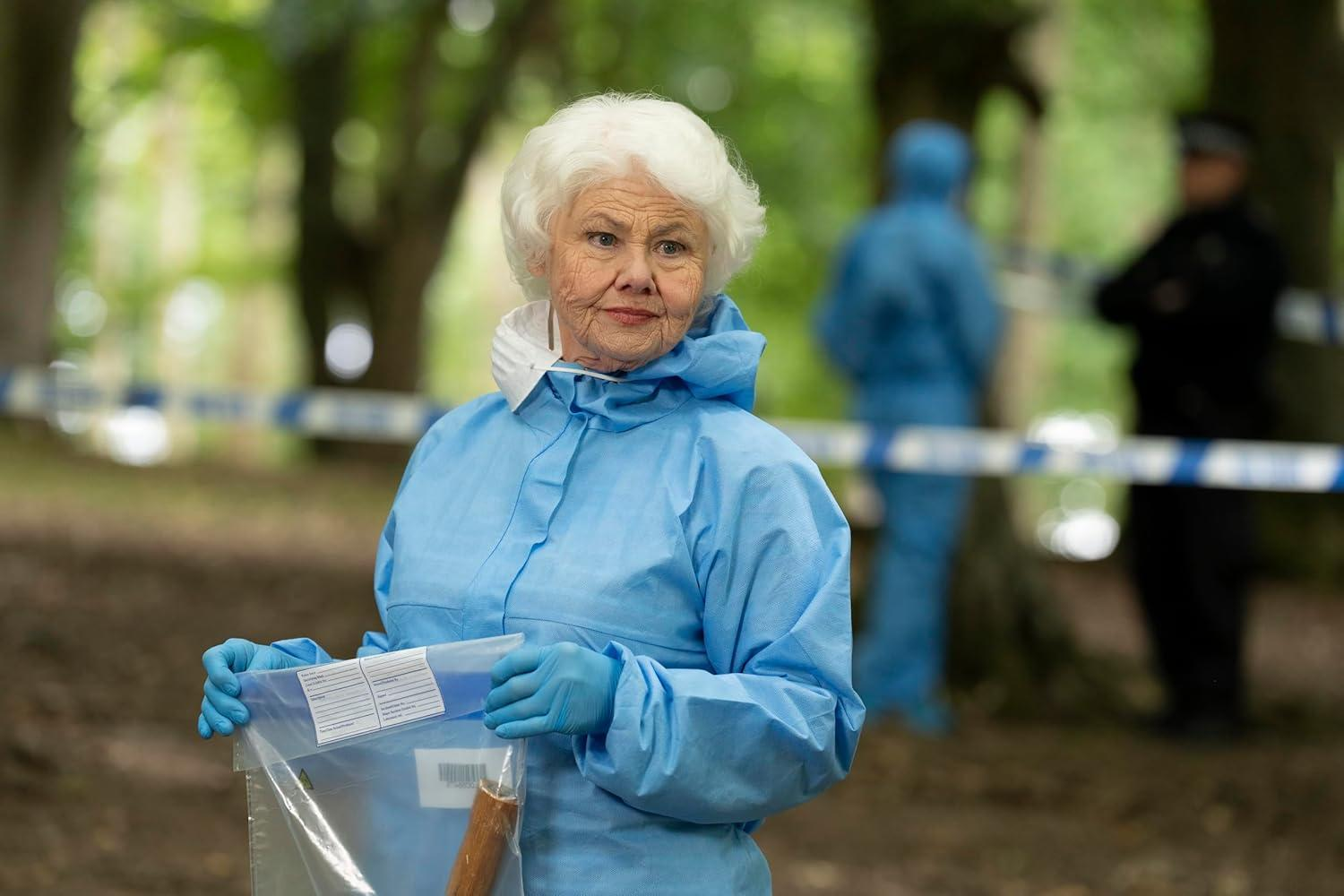 Eine ältere Frau in einem blauen forensischen Anzug hält eine Tüte mit Beweismitteln. Im Hintergrund sind zwei Figuren und Absperrband zu sehen.
