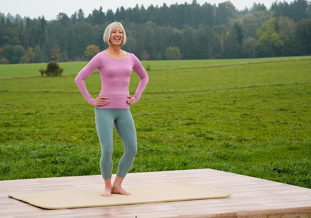 Eine ältere Frau in Sportkleidung lächelt auf einer Yogamatte. Grünes Feld und Bäume sind im Hintergrund.