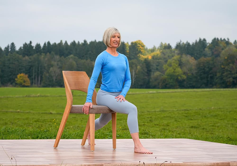Frau in blauem Oberteil und grauer Leggings macht Yoga auf einem Stuhl im Freien. Grünes Feld und Bäume im Hintergrund.