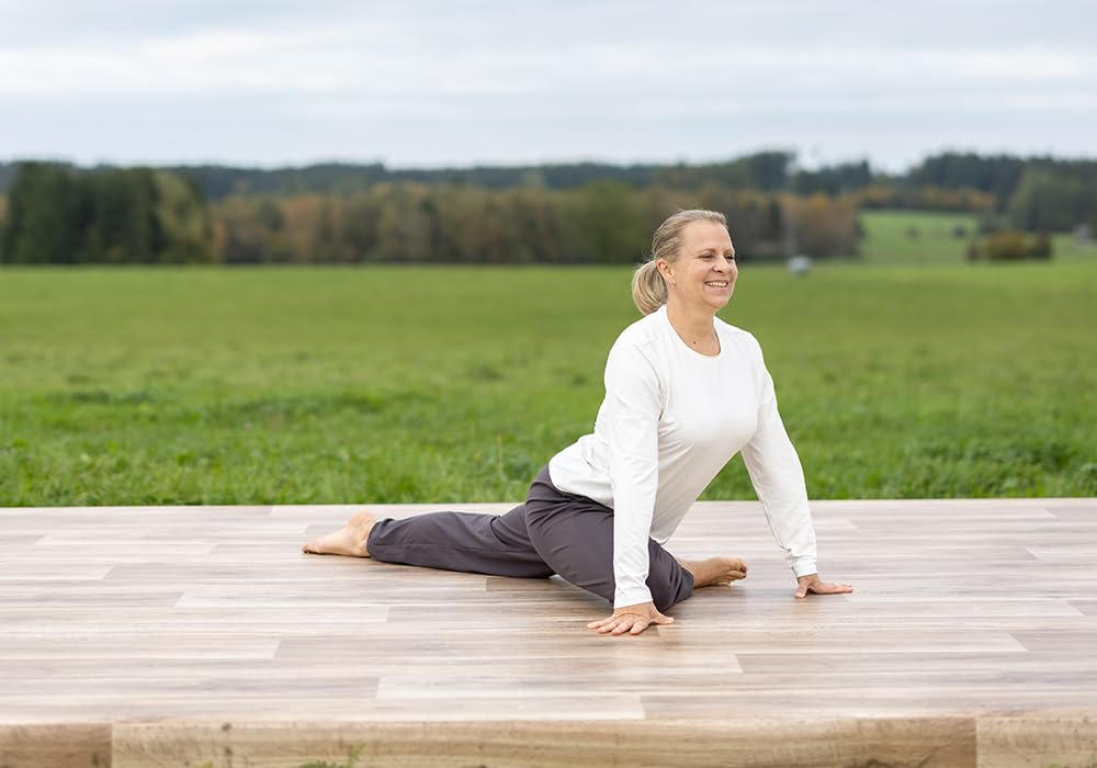 Frau in Yoga-Pose auf Holzplattform. Grünes Feld und Bäume im Hintergrund. Sie trägt ein weißes Oberteil und graue Hose.