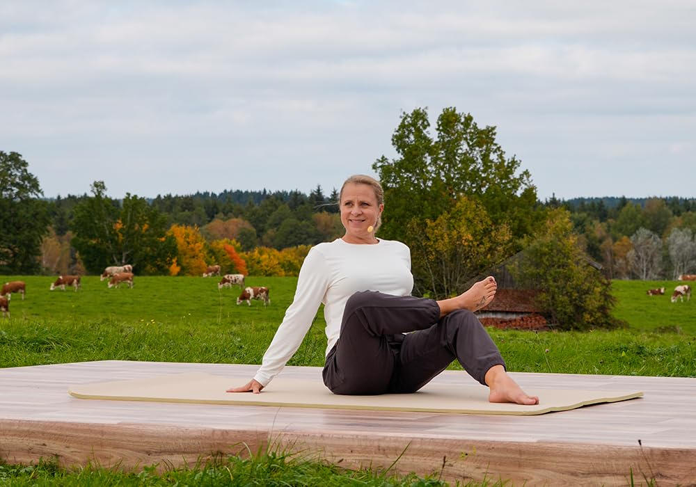 Frau in Yoga-Pose auf einer Matte. Kühe grasen auf einer grünen Wiese hinter ihr. Der Himmel ist bedeckt.