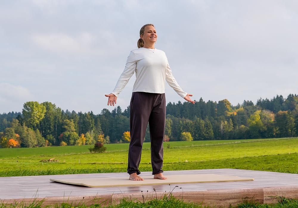 Frau in weißem Oberteil und schwarzer Hose steht auf einer Matte, Arme offen. Grünes Feld und Bäume im Hintergrund.