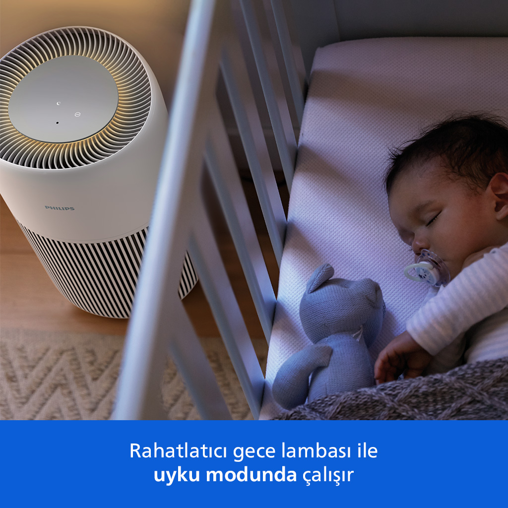 Bir bebek beşiğinde uyuyor, bir oyuncak ayının yanında. Arka planda bir hava temizleyici var.