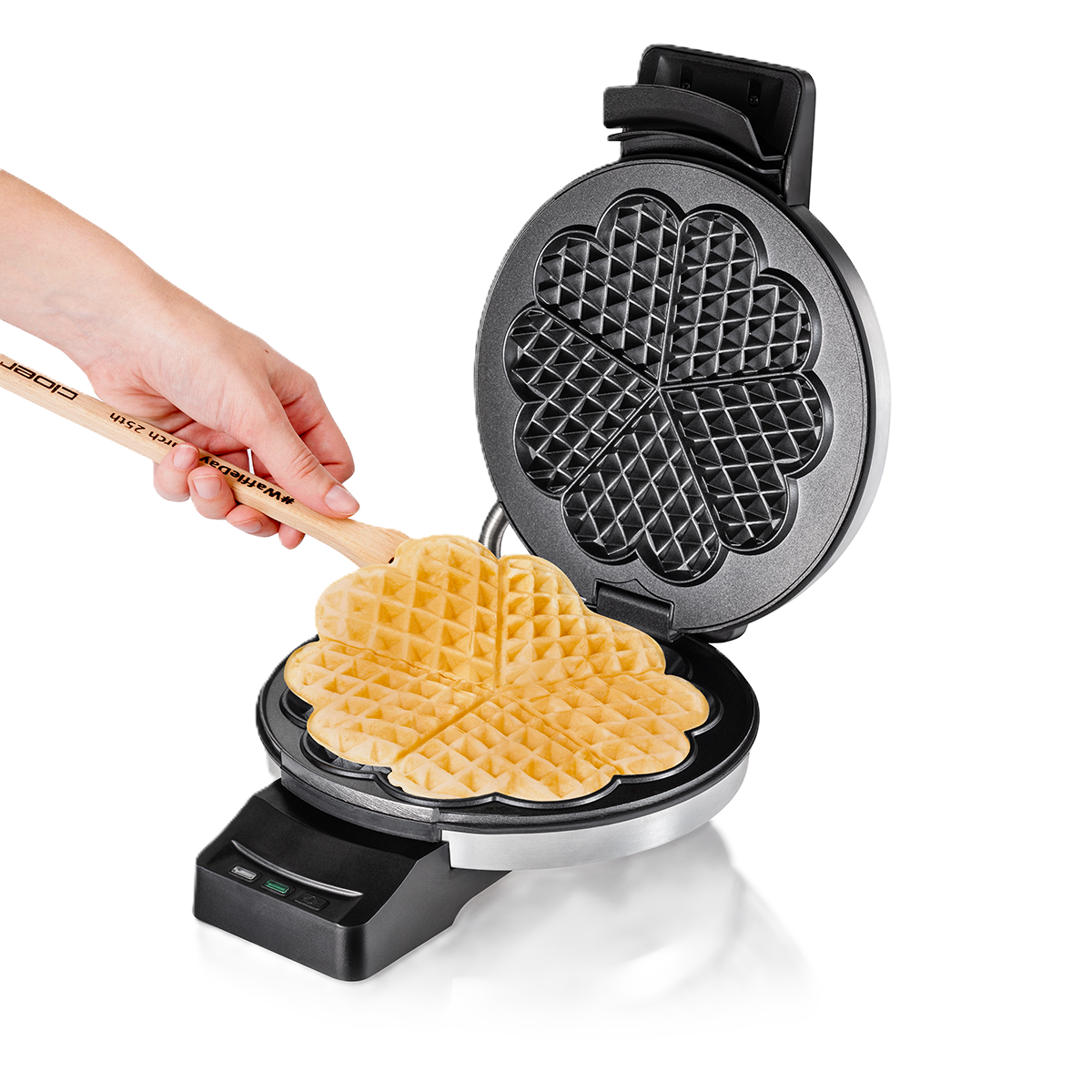 Eine Hand hält einen Holzspatel und hebt einen herzförmigen Waffel aus einem offenen Waffeleisen. Die Waffel ist goldbraun.