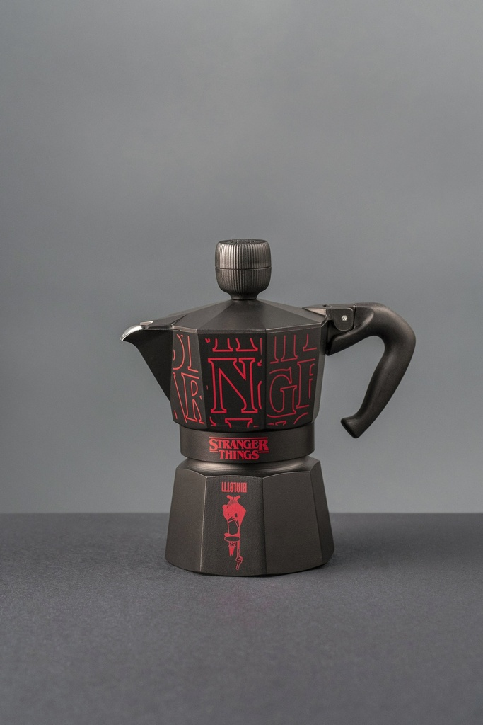 Schwarze Kaffeemaschine mit rotem Aufdruck Stranger Things. Auf dunkler Oberfläche, grauer Hintergrund.