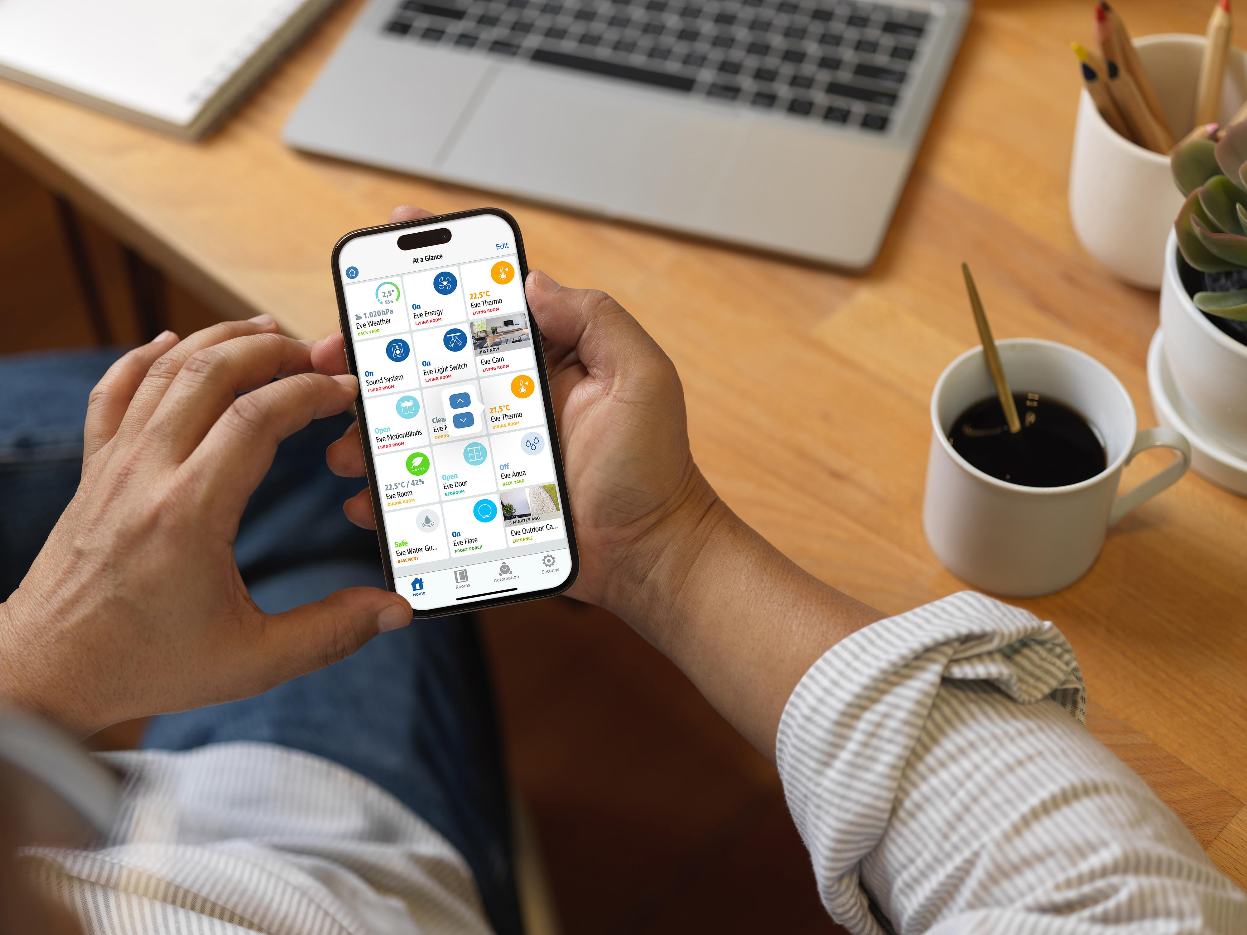Eine Person hält ein Telefon mit einer Smart-Home-App. Laptop, Kaffee und Pflanzen sind im Hintergrund zu sehen.